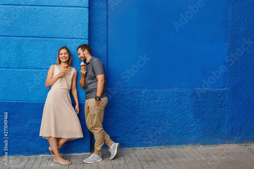 Happy couple having fun, eating ice cream and smiling against the blue wall
