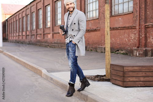 Handsome young man in grey coat and hat crossing the street with a cup of coffee.