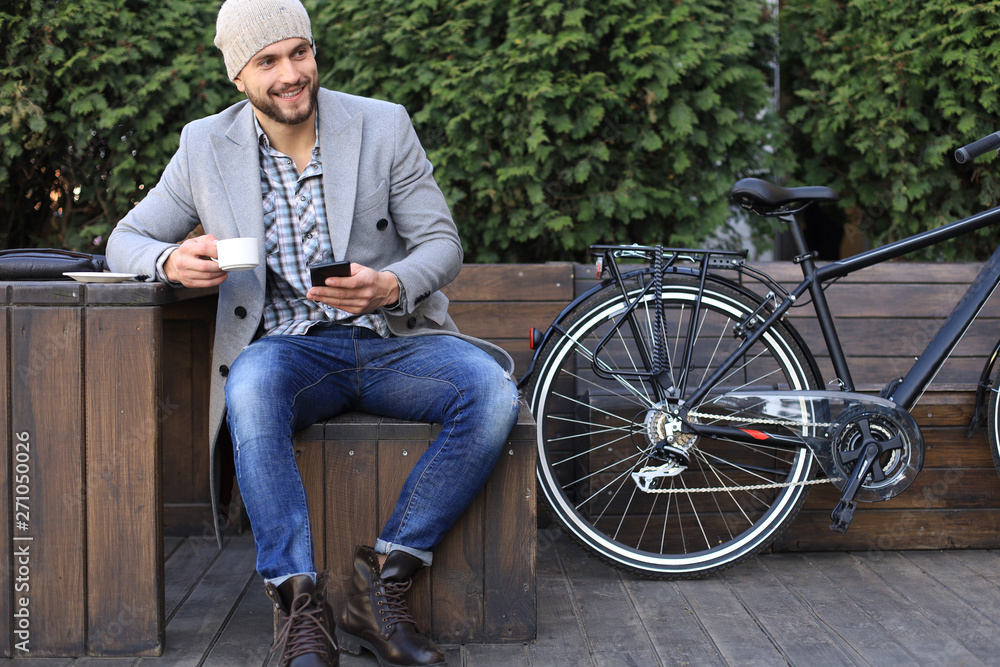 Obraz premium Handsome young man in grey coat and hat using smartphone, resting, and smiling while sitting near his bicycle outdoors.