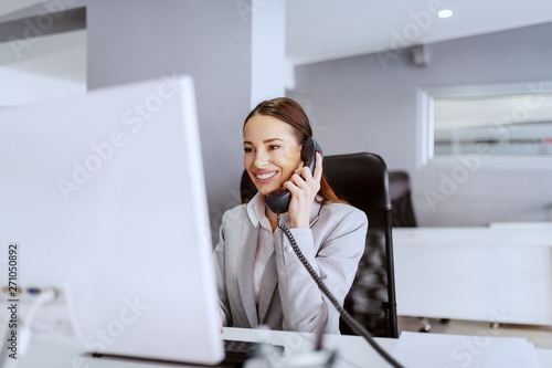 Gorgeous Caucasian businesswoman with long brown hair and in formal wear using computer and talking on the phone. Multitasking concept. photo