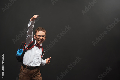 joyful little boy, on a black background with a briefcase behind his shoulders shows a gesture of victory, joy of success. Happy emotional child pupil rejoices back to school.