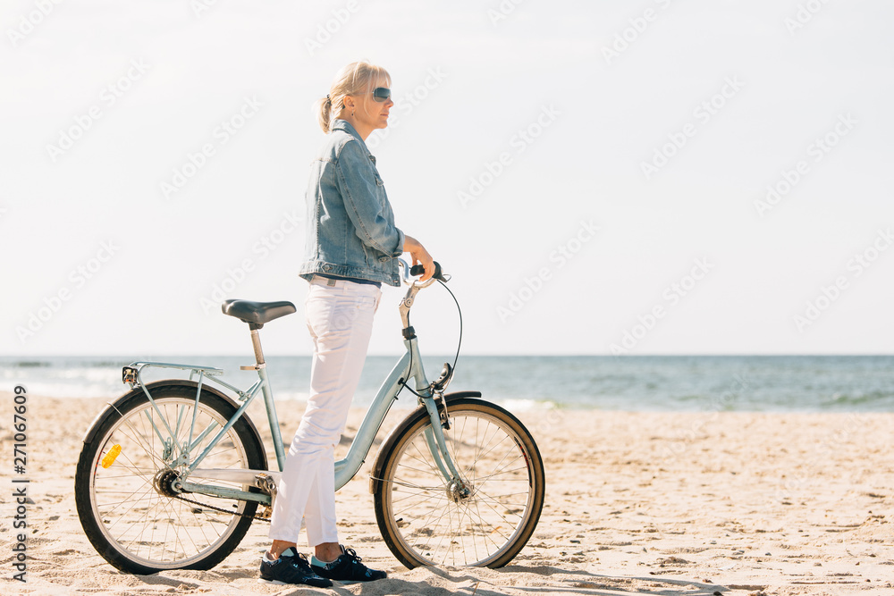 Fototapeta premium Pretty blonde girl in white pants and denim coat standing on the beach with bicycle. Atrractive woman relaxing near the sea after bike ride