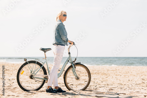 Pretty blonde girl in white pants and denim coat standing on the beach with bicycle. Atrractive woman relaxing near the sea after bike ride © beatleoff