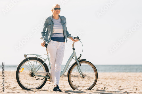 Pretty blonde girl in white pants and denim coat standing on the beach with bicycle. Atrractive woman relaxing near the sea after bike ride