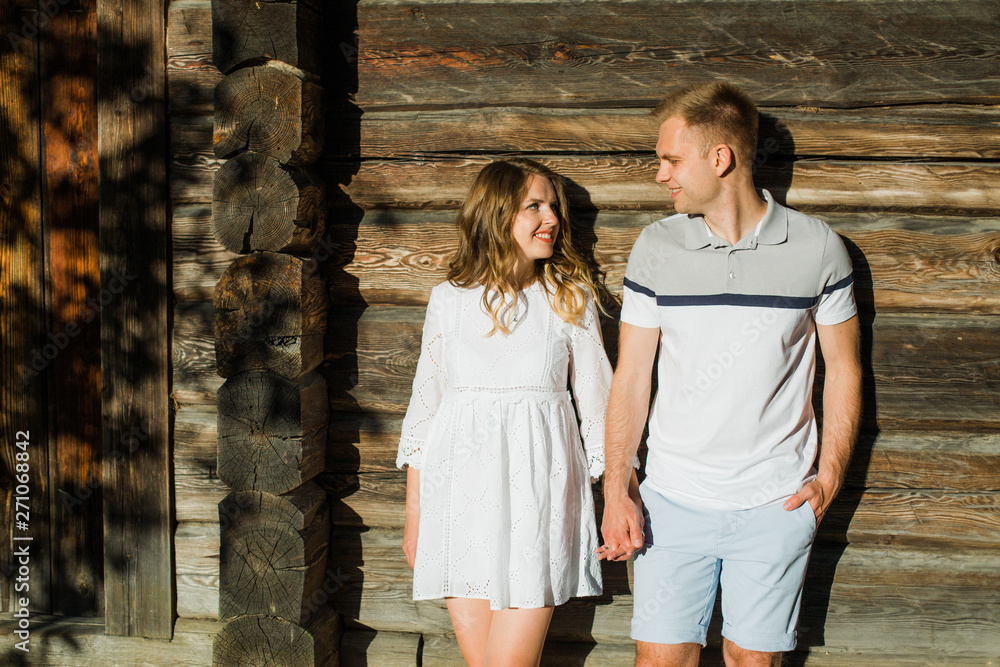 Young pretty couple in love standing near wooden house. Handsome cheerful blonde girl in white dress with her boyfriend. Man and woman having fun outdoors
