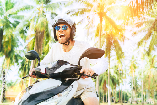 Happy smiling and screaming male tourist in helmet and sunglasses riding motorbike scooter during his tropical vacation under palm trees