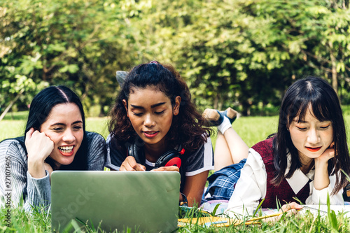 Group of smiling international students or teenagers sitting and using laptop computer doing their homework with book together in park at university.Education and friendship Concept