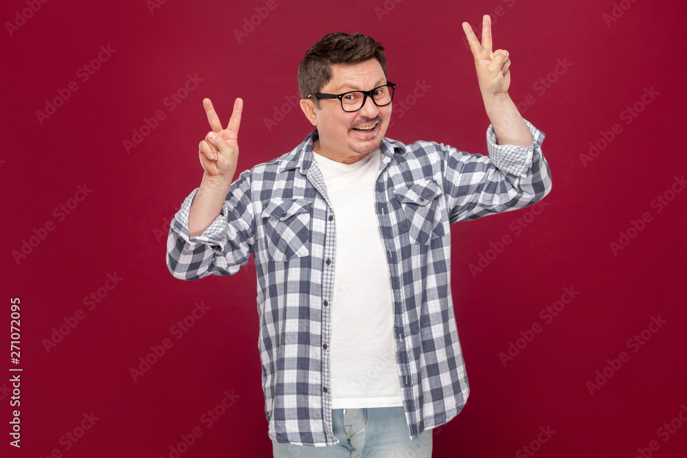 Portrait of funny happy middle aged man in casual checkered shirt, eyeglasses standing with peace or victory sign and looking at camera with toothy smile. studio shot, isolated on dark red background. Portrait of funny happy middle aged man in casual checkered shirt, eyeglasses standing with peace or victory sign and looking at camera with toothy smile. studio shot, isolated on dark red background.