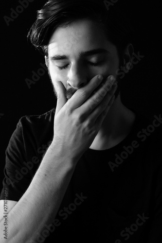 Close up of young adult male eyes closed hand over mouth in shock or horror. Monotone, black and white for dramatic effect, dark and moody series. Concept image for distress or realization of actions.