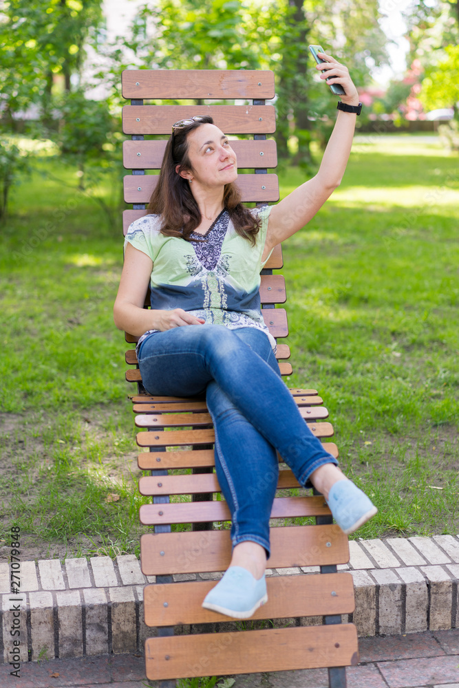 Naklejka premium Young woman making selfie in the park. Sexy young woman with long blond hair doing selfie on summer day in park. vertical photo