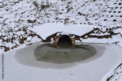 Closeup photograph of a drain pipe that provides storm water to a wet retention basin. The retention basin is partially covered in a thin layer of ice. Snow covers the retention basin's shore. 