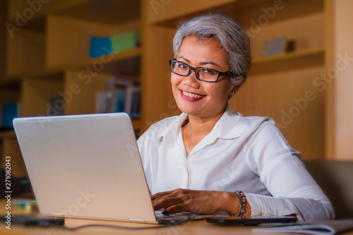 Corporate portrait of attractive and happy successful mature Asian woman working at laptop computer desk smiling confident and charming
