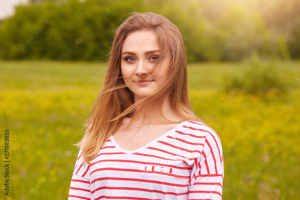 Close up portrait of beautiful woman with fair hair enjoying nature, relaxing in open air. Lady posing in green grass meadow, wearing white shirt with red srtipes, looks at camera. Tenderness concept.
