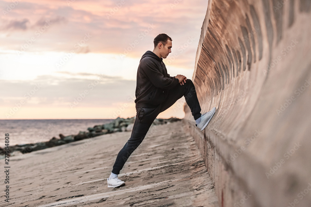 Slim handsome young guy fitness coach doing stretching for muscles and leg bundles before jogging on a rocky beach against background of sea and setting sun on a warm summer evening. Advertising space
