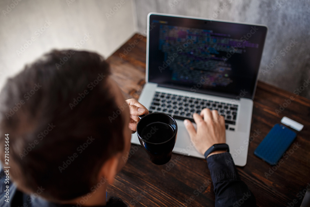 a male programmer in a black shirt is working at a laptop, close-up of the program code and developer's hands on a laptop. modern young programmer sitting at a wooden table at a computer a male programmer in a black shirt is working at a laptop, close-up of the program code and developer's hands on a laptop. modern young programmer sitting at a wooden table at a computer