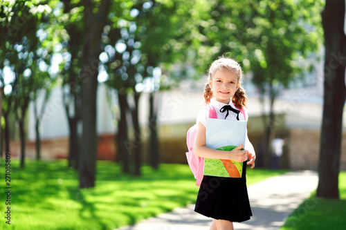 Smiling student girl wearing school backpack and holding exercise book. Portrait of happy Caucasian young girl outside the primary school. Closeup face of smiling schoolgirl looking at camera.