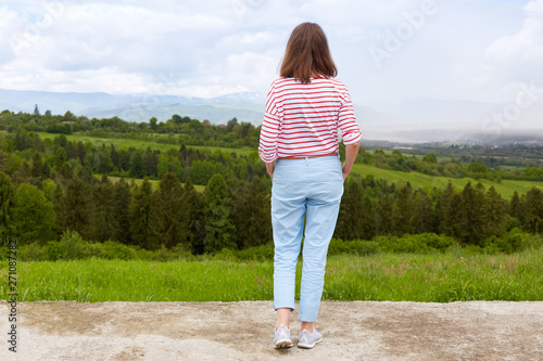 Woman standing in mountain, panoramic view of mountain range, trees. green grass. blue sky, female wearing casual trousers and white t shirt with red stripes enjoing beautiful nature. People concept. © sementsova321