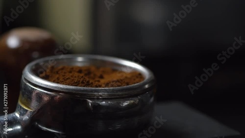 Close-up hand shot of barista putting portafilter down on tamping stand, pressing freshly ground coffee with tamper, then removing portafilter from frame, against dark blurred background