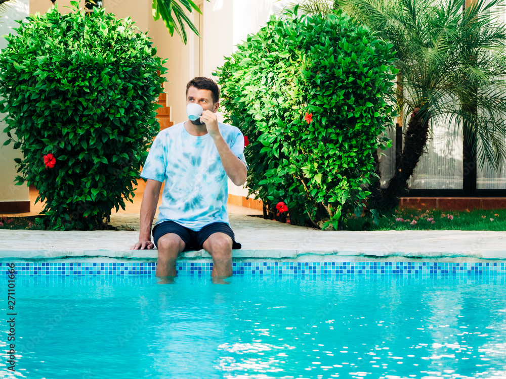 Portrait of a mature age man wearing a t-shirt and shorts sitting near a swimming pool drinking coffee. feet in the pool in the water.