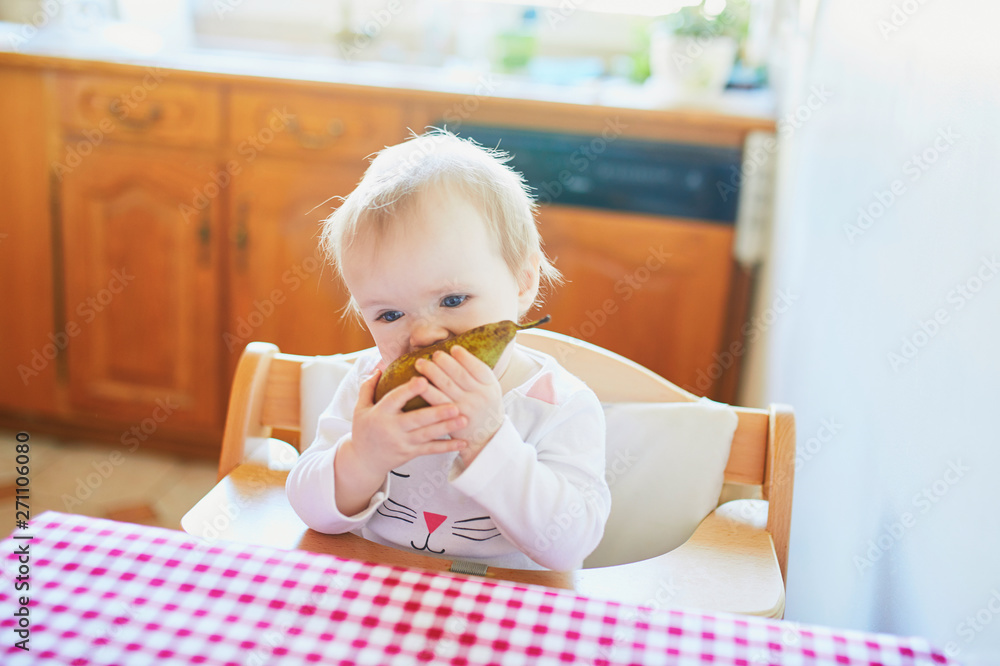 Fototapeta premium Cute baby girl eating pear in the kitchen
