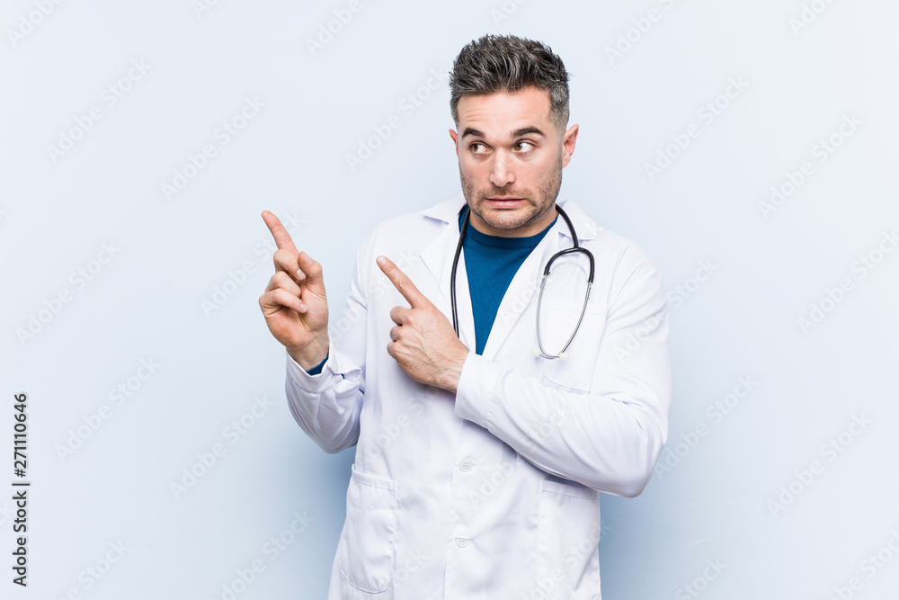 Young handsome doctor man shocked pointing with index fingers to a copy space. Young handsome doctor man shocked pointing with index fingers to a copy space.
