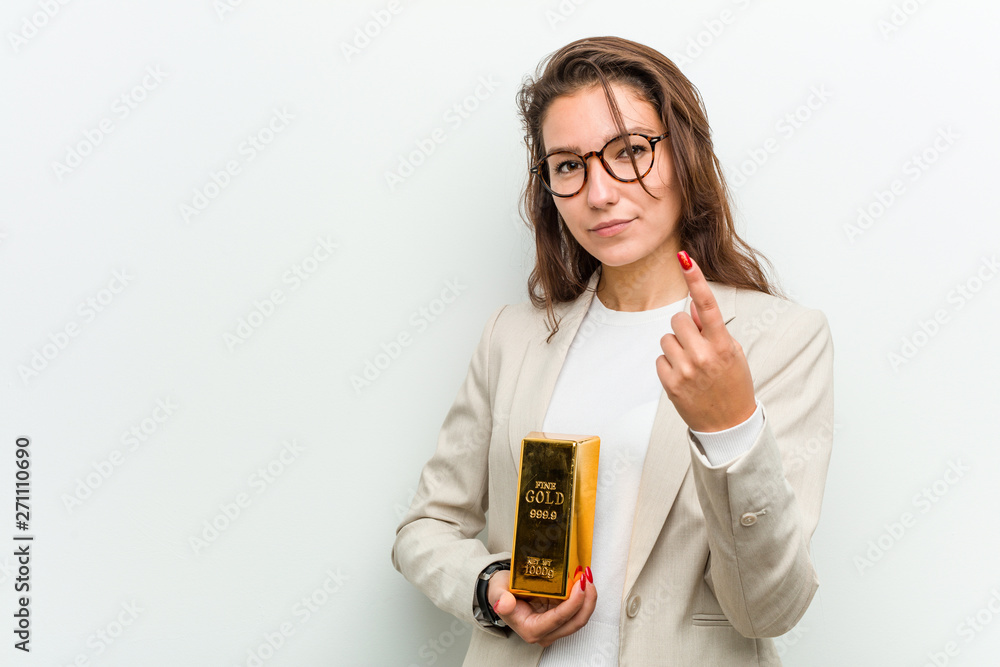 Young european business woman holding a gold ingot pointing with finger at you as if inviting come closer. Young european business woman holding a gold ingot pointing with finger at you as if inviting come closer.