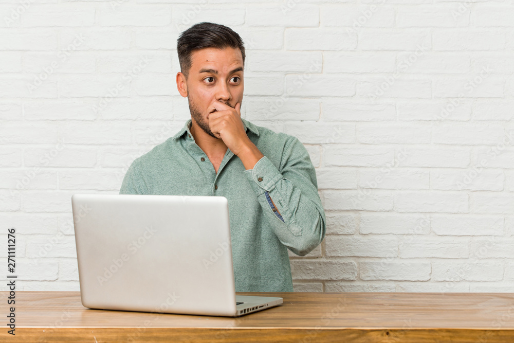 Young filipino man sitting working with his laptop shocked pointing with index fingers to a copy space. Young filipino man sitting working with his laptop shocked pointing with index fingers to a copy space.