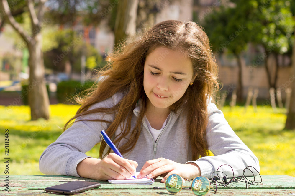 Cute teen girl writes notes on a paper pad by wooden table on a green meadow. Cute teen girl writes notes on a paper pad by wooden table on a green meadow.