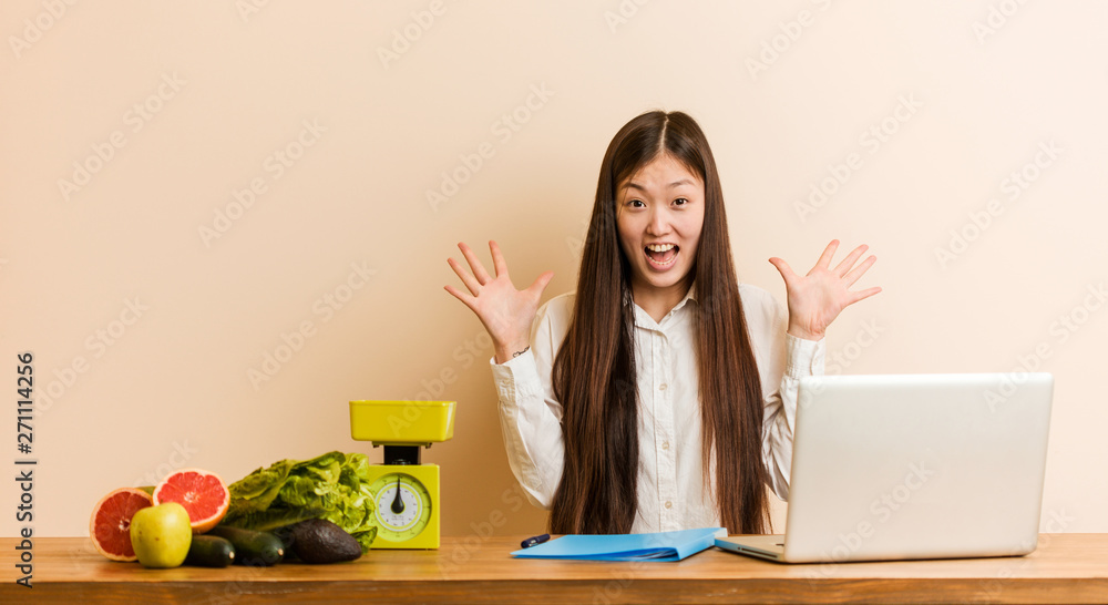 Young nutritionist chinese woman working with her laptop celebrating a victory or success Young nutritionist chinese woman working with her laptop celebrating a victory or success