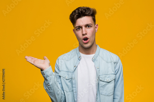 Young handsome student wearing a denim shirt impressed holding copy space on palm.