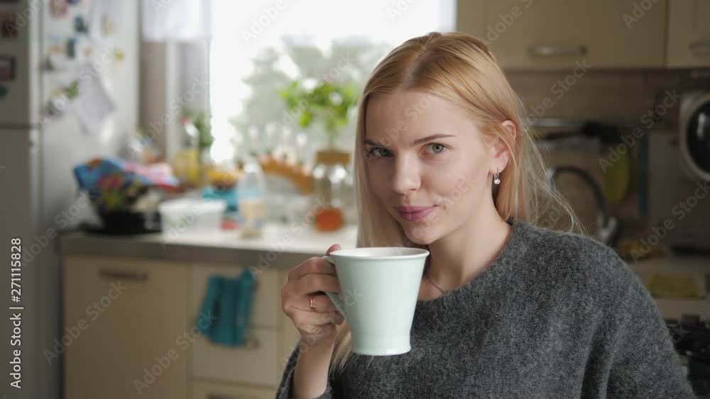 Medium closeup shot of young smiling adorable blonde european woman in grey sweater with mug posing against blurred cozy domestic kitchen interior background, looking out the window, smiling, laughing Medium closeup shot of young smiling adorable blonde european woman in grey sweater with mug posing against blurred cozy domestic kitchen interior background, looking out the window, smiling, laughing
