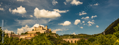 View of Spoleto, green mountains, blue sky with white clouds. The Rocca Albornoziana fortress illuminated by the sun in summer. The bridge of the towers, Roman aqueduct. Trees in the foreground photo