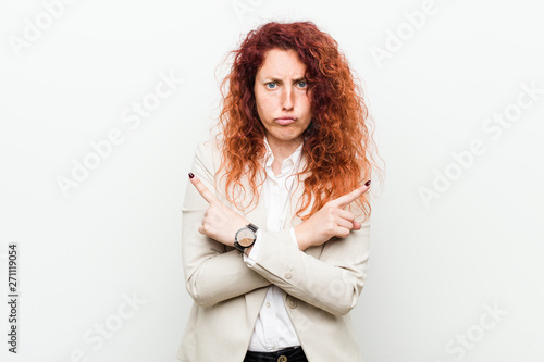 Young natural redhead business woman isolated against white background points sideways, is trying to choose between two options.