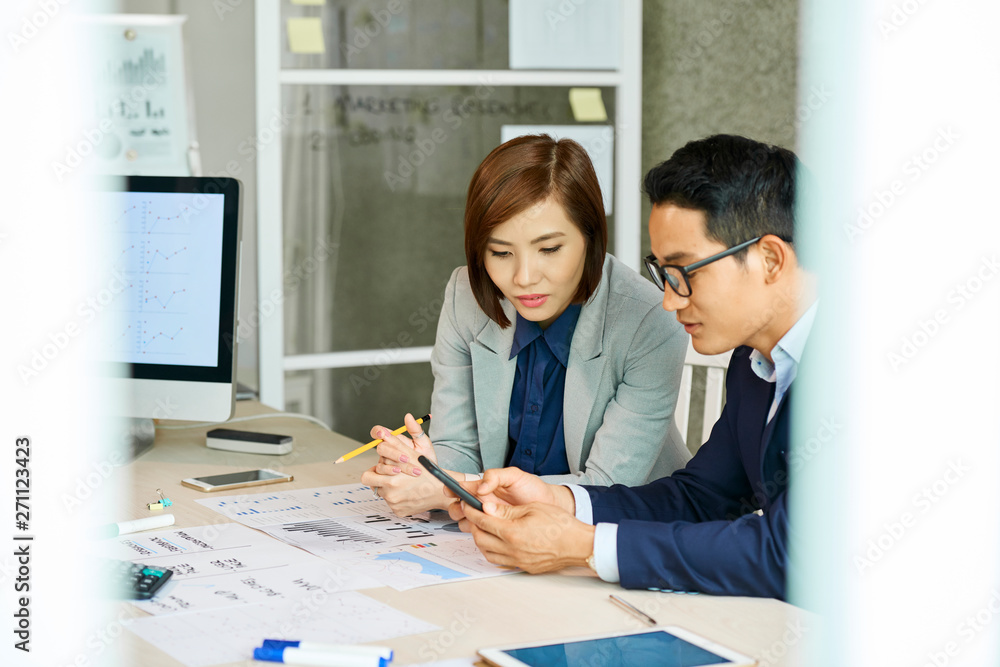 Vietnamese businessman and businesswoman sitting at office table with various documents and reading text message Vietnamese businessman and businesswoman sitting at office table with various documents and reading text message
