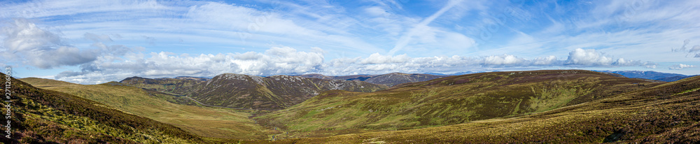 A panorama view of a Scottish mountain valley with heather, road and mountain range under a majestic blue sky and white clouds