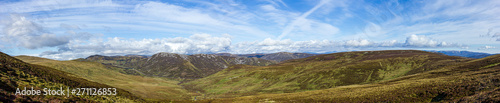 A panorama view of a Scottish mountain valley with heather, road and mountain range under a majestic blue sky and white clouds