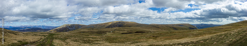 A panorama view of a Scottish mountain summit plateau with heather and path trail under a majestic blue sky and huge white clouds