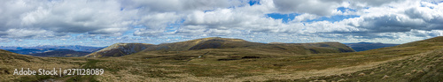 A panorama view of a Scottish mountain summit plateau with heather and path trail under a majestic blue sky and huge white clouds