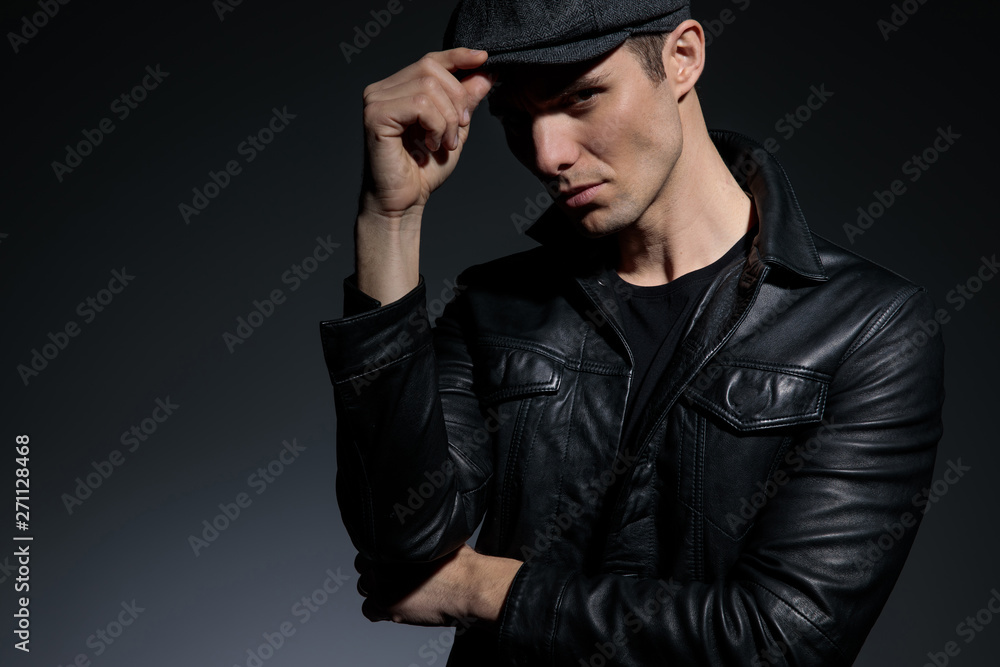 Close up a serious looking man adjusting his cap and confidently staring to the camera while wearing a black leather jacket and standing on white studio background