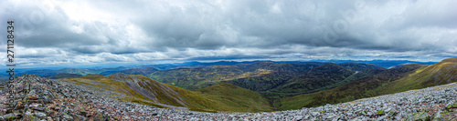 A panorama view of a Scottish mountain valley with a road and mountain range under a stormy huge white clouds sky