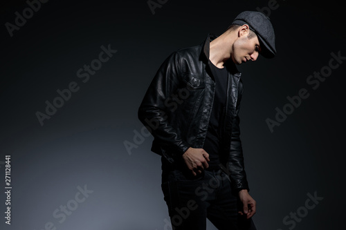 Thoughtful man looking down upset and holding his hand on his leather jacket while wearing a hat and black jeans, posing on gray studio background