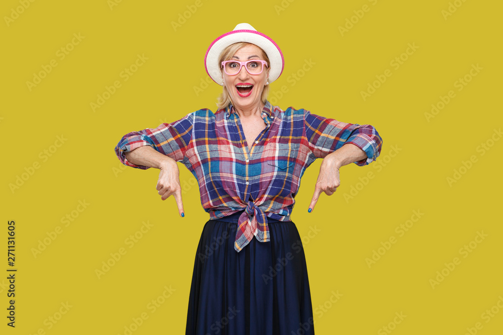 Portrait of surprised modern stylish mature woman in casual style with hat and eyeglasses standing, amazed looking at camera and pointing down. indoor studio shot isolated on yellow background. Portrait of surprised modern stylish mature woman in casual style with hat and eyeglasses standing, amazed looking at camera and pointing down. indoor studio shot isolated on yellow background.