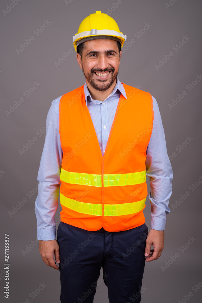 Handsome bearded Persian man construction worker against gray ba