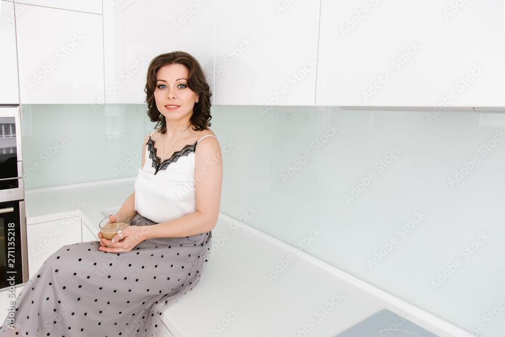 A young beautiful Caucasian woman sits at a table in the white modern kitchen, the housewife drinks coffee with milk. A young beautiful Caucasian woman sits at a table in the white modern kitchen, the housewife drinks coffee with milk.