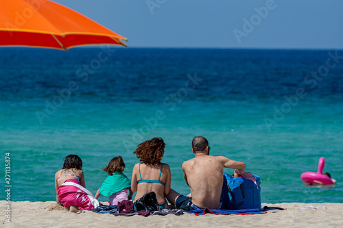 Unidentified family with two children sitting back on white sandy beach and looking to crystal clear blue sea water.