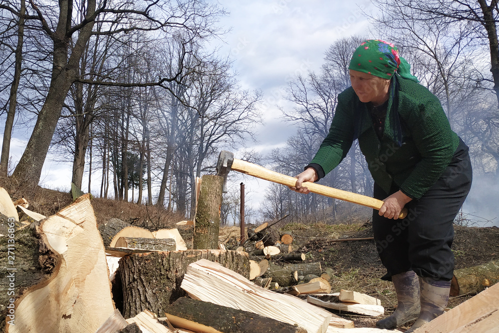 An old woman chopping firewood for the winter, also removes her yard near the house, on the street early spring. An old woman chopping firewood for the winter, also removes her yard near the house, on the street early spring.