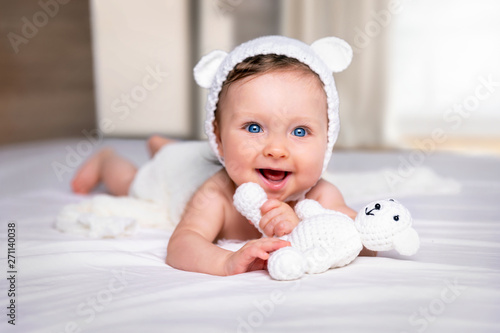 Portrait eines glücklichen, süßen Baby Mädchens mit blauen Augen und Bärenmütze auf einem weißem Bett mit Stoff Bär photo