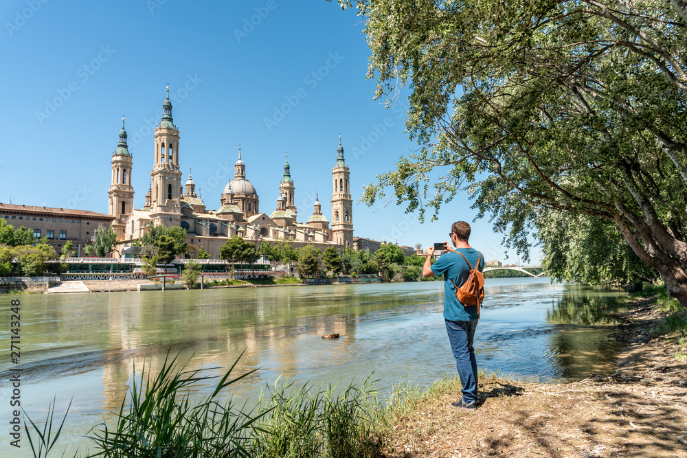 Fototapeta premium Male tourist standing at the Ebro river bank and taking a photo of El Pilar in sunny day. 