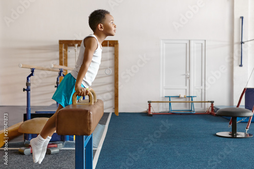 Horizontal shot of skillful atheltic Afro American boy in sportswear mounting on pommel horse, keeping hands on handles, performing difficult gymnastic exercise at gym. Childhood, ethnicity and sports