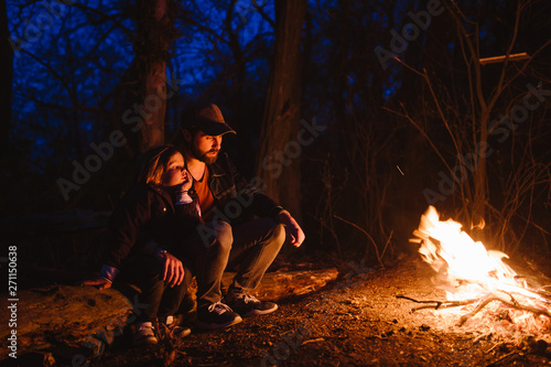 Father and his little son sitting together on the logs in front of a fire at the night. The hike in the forest.